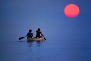Two people paddling a canoe in deep blue water with a strong red sun in the background