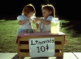 Two little girld selling lemonade at a stand while sampling their wares